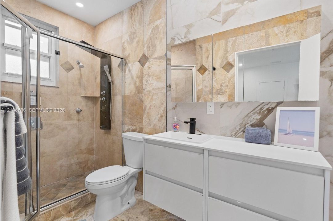Full-wall stone tiling, frameless glass shower enclosure, matte black fixtures, white dual-drawer vanity with integrated sink, and mirrored medicine cabinet