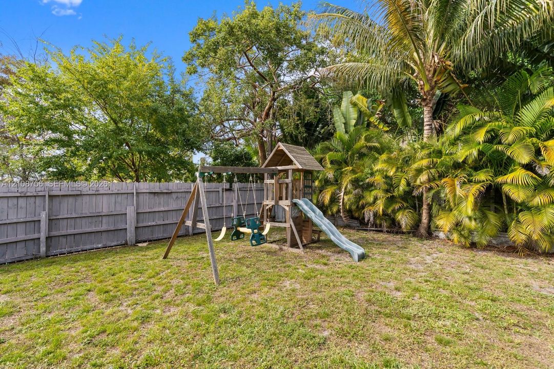 Fenced backyard featuring mature trees, tropical palms, and a wooden play structure