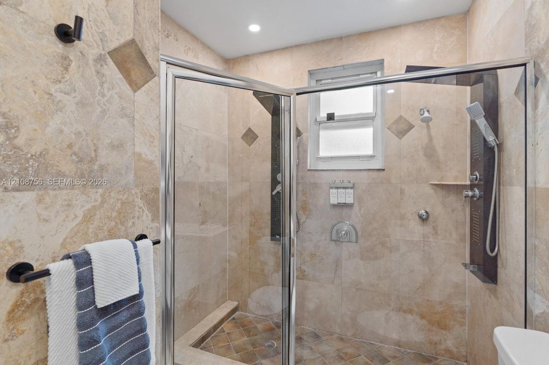 Walk-in shower featuring natural stone wall tile, decorative accent tiles, a multi-functional shower panel system with a handheld sprayer, and a glass enclosure