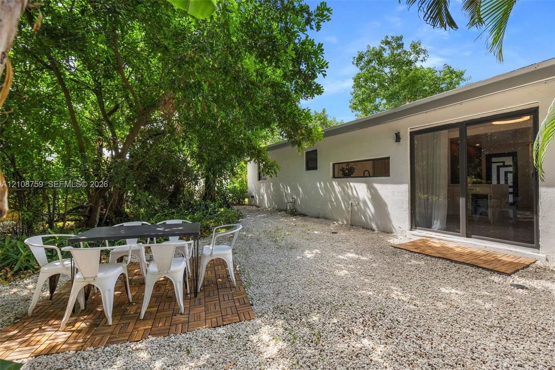 Exterior space featuring light-colored stucco siding, gravel ground covering, sliding glass doors, and mature trees providing shade
