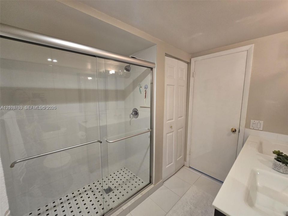 Walk-in shower with sliding glass doors, white subway-style wall tiling, mosaic floor tile, dual-basin vanity with a solid-surface countertop, and double-door closet storage