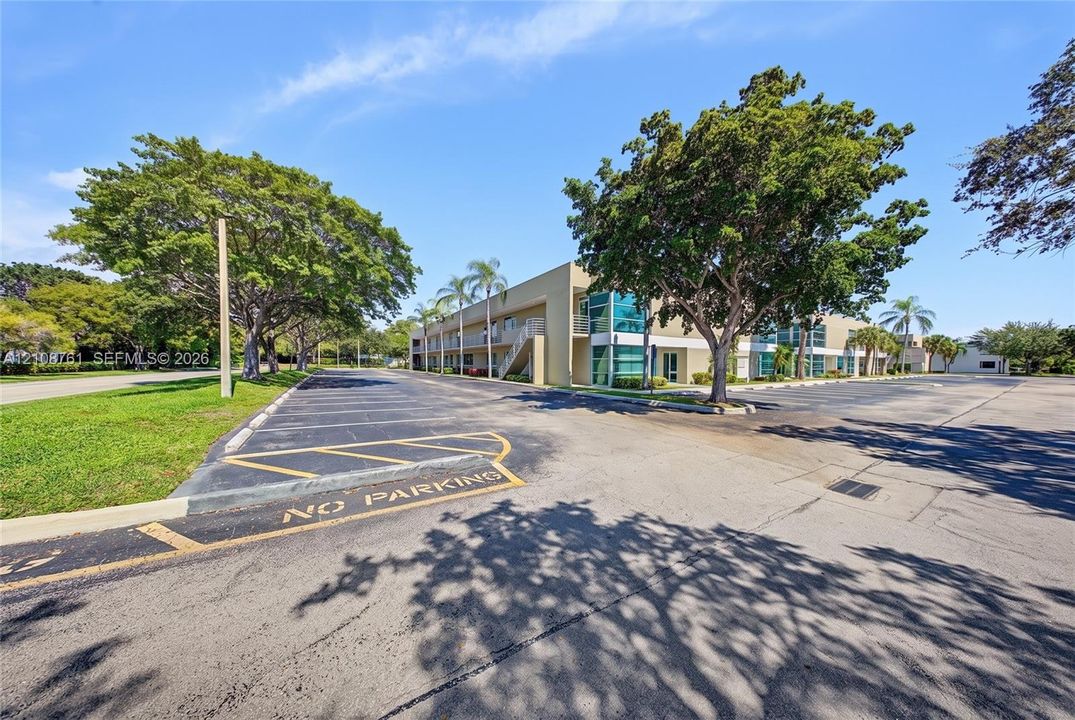 Multi-story exterior featuring a flat roofline, glass paneling, and an expansive paved parking surface surrounded by mature trees