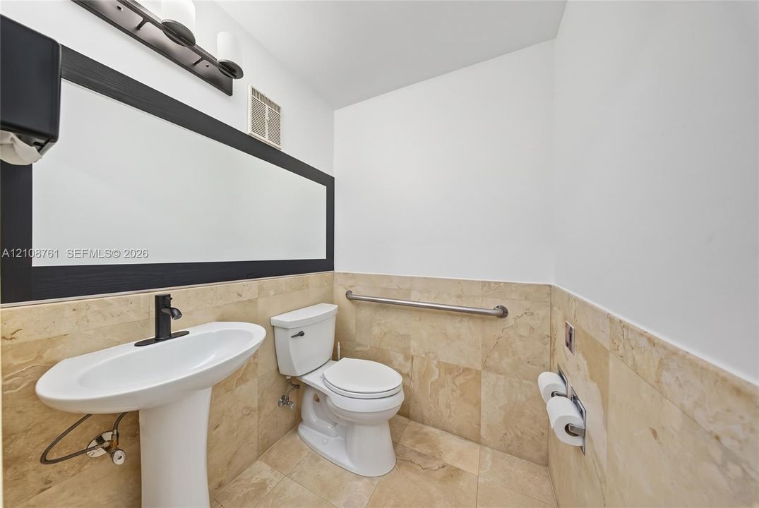 Pedestal sink with matte black faucet, wall-mounted mirror with black frame, stone wall and floor tiling, and stainless steel grab bar