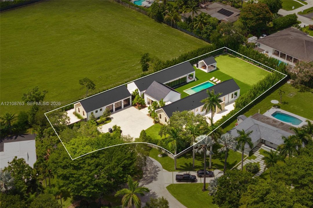Estate featuring a multi-structure layout with metal roofing, a swimming pool, detached gazebo, expansive paved motor court, and manicured grounds enclosed by privacy hedges