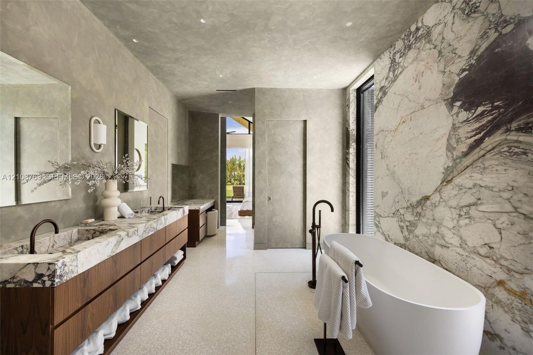 Custom vanity with integrated stone sinks and wood-finish cabinetry, freestanding soaking tub, expansive stone-slab feature wall, and recessed ceiling lighting