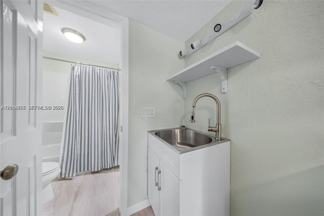 White vanity featuring a stainless steel drop-in sink, high-arc gooseneck faucet, and wall-mounted shelving, paired with a multi-light fixture