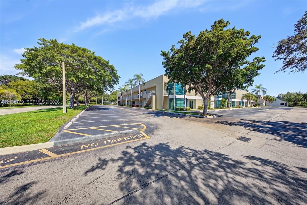 Multi-story exterior featuring a flat roofline, glass paneling, and an expansive paved parking surface surrounded by mature trees