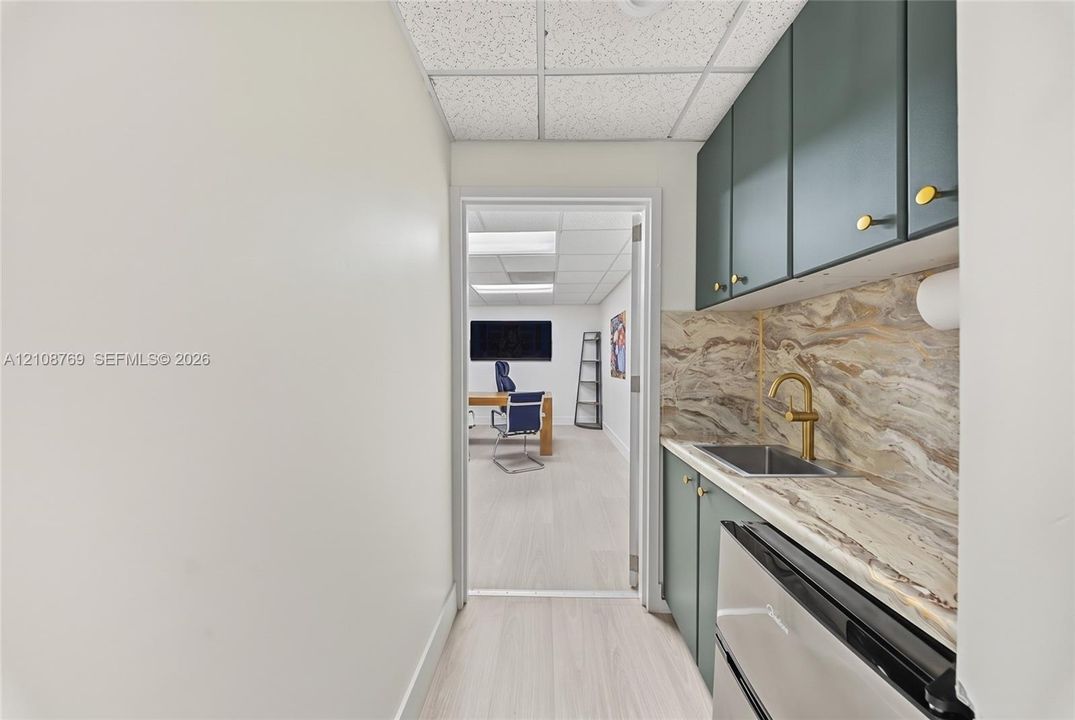 Kitchenette featuring custom dark cabinetry, stone-patterned backsplash, undermount stainless steel sink, gold-tone hardware, and light-toned wood-finish flooring