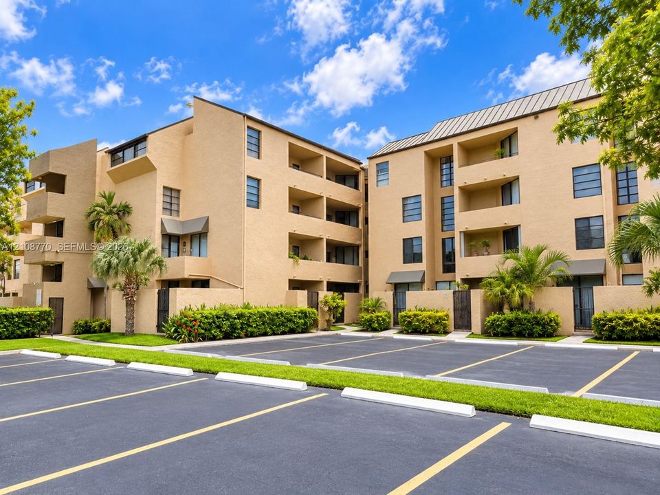 Multi-story stucco exterior featuring private recessed balconies, dark-framed windows, metal roofing, and asphalt parking area