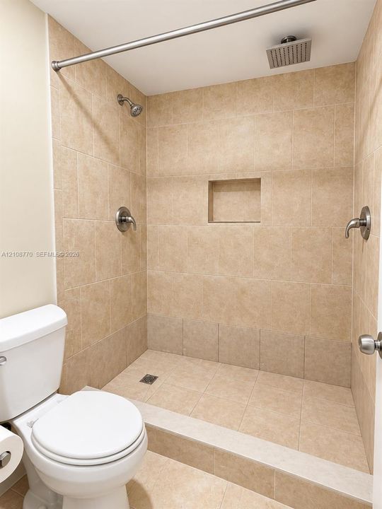 Walk-in shower featuring floor-to-ceiling neutral tile, built-in recessed storage niche, dual showerheads including an overhead rainfall fixture, and a stone-surface threshold