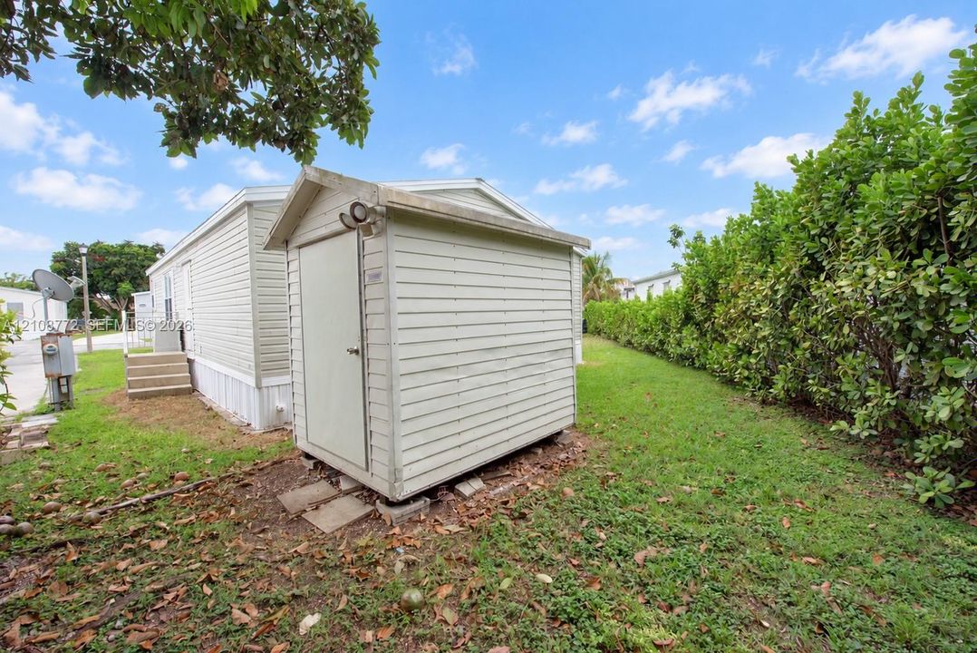Exterior storage shed with horizontal siding, set on a grass-covered lot bordered by mature privacy hedges