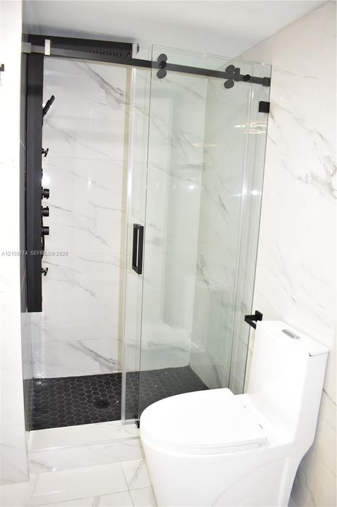 Walk-in shower featuring a frameless sliding glass door, matte black hardware, multi-setting shower panel, and hexagonal floor tiling, complemented by white large-format wall tile and a dual-flush toilet