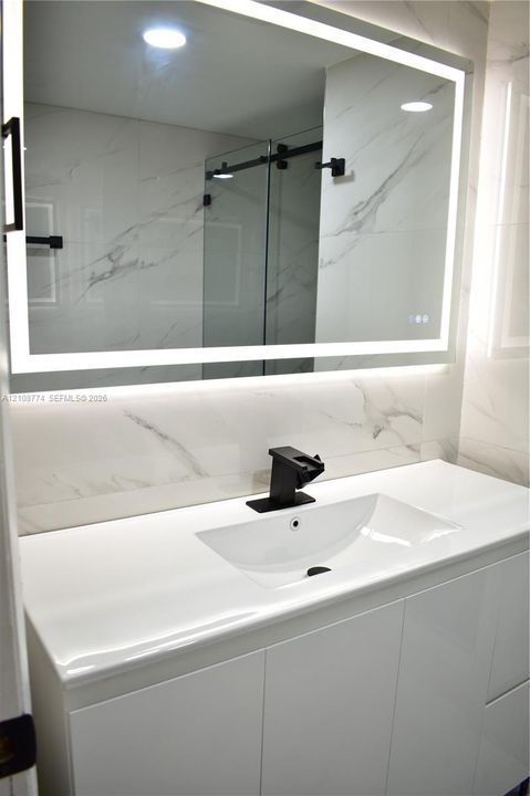 Modern vanity with a white countertop and integrated sink, matte black faucet, wall-mounted LED-backlit mirror, and stone-finish wall tile