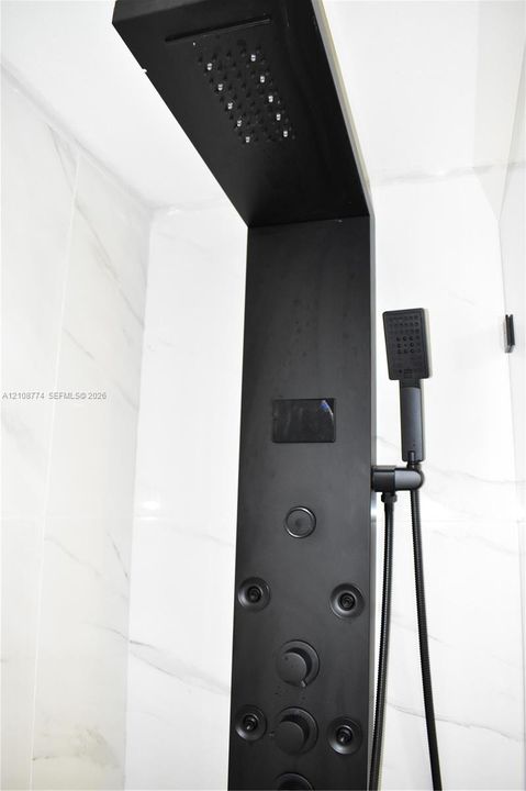 Matte black shower panel featuring an integrated rainfall showerhead, handheld sprayer, body jets, and a digital temperature display against large-format, light-toned stone-finish wall tile