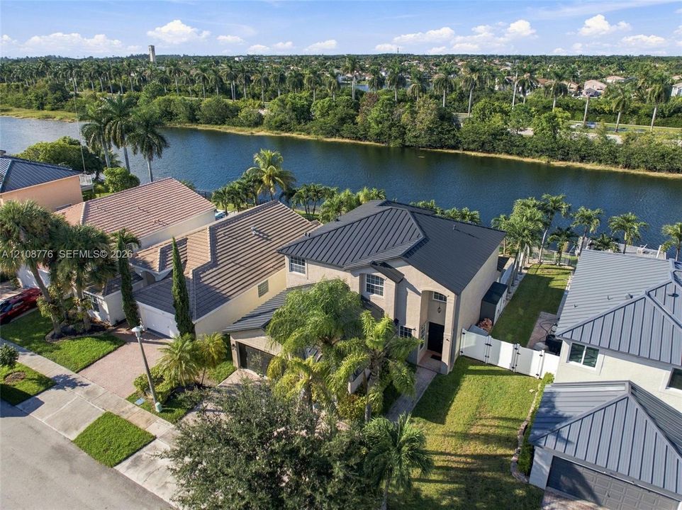 Stucco exterior residence featuring a metal roof, paved driveway, white vinyl perimeter fencing, and water frontage