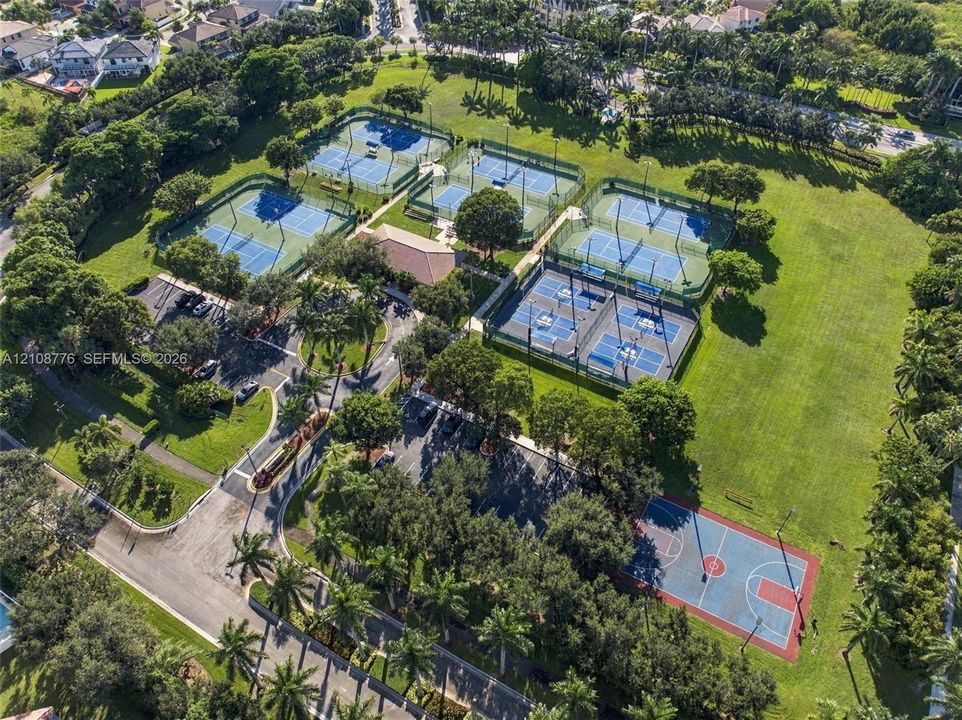 Recreational complex featuring multiple tennis courts, a paved basketball court, asphalt parking areas, and mature trees situated on expansive lawn grounds