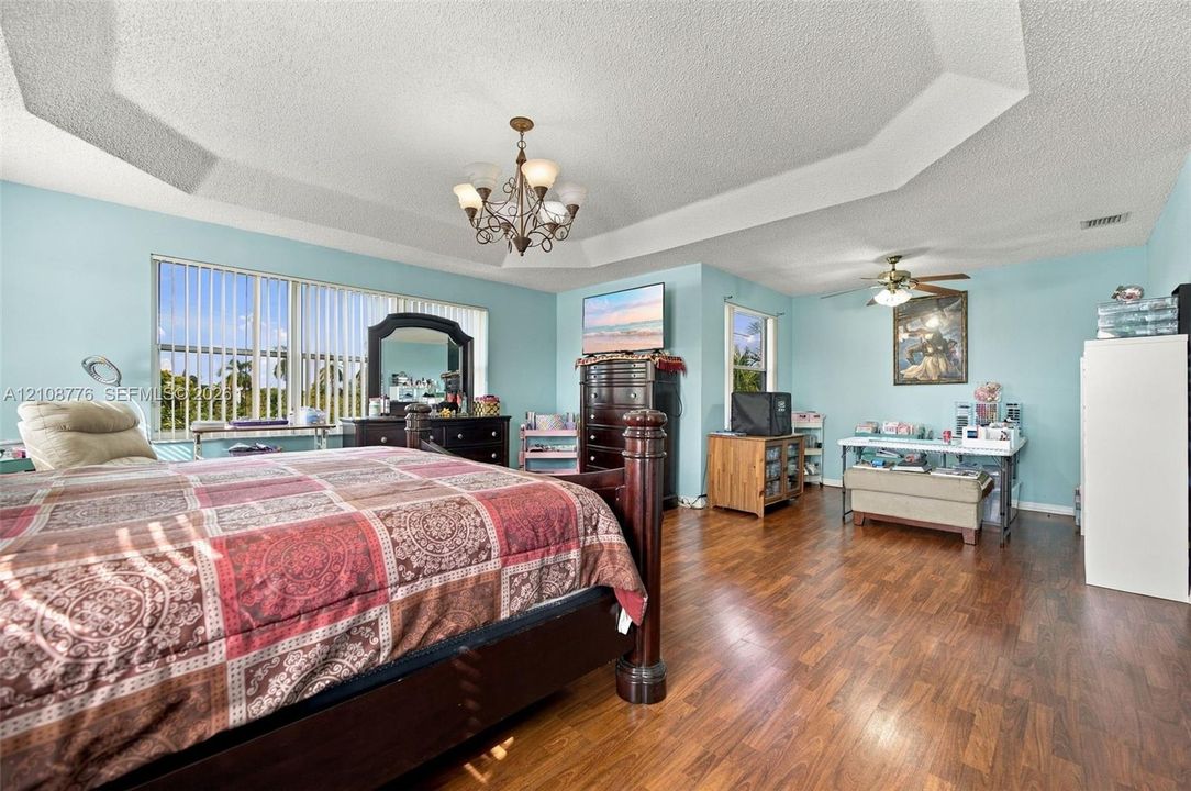 Tray ceiling with recessed lighting, wood-finish flooring, multi-pane window with vertical blinds, and a central tiered chandelier