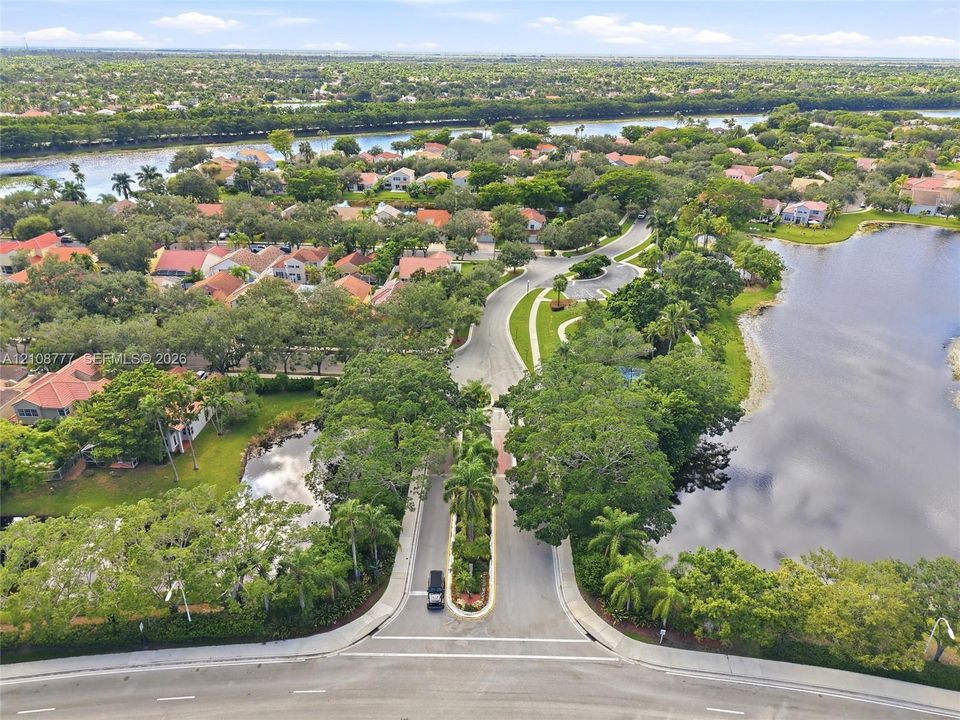 Aerial perspective of a community entrance featuring paved roadways, mature tree canopy, waterfront borders, and landscaped median