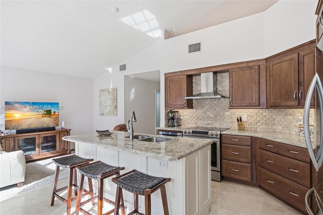 Kitchen featuring custom wood cabinetry, stone countertops, stone-tile backsplash, stainless steel range hood, and a central island with a built-in sink