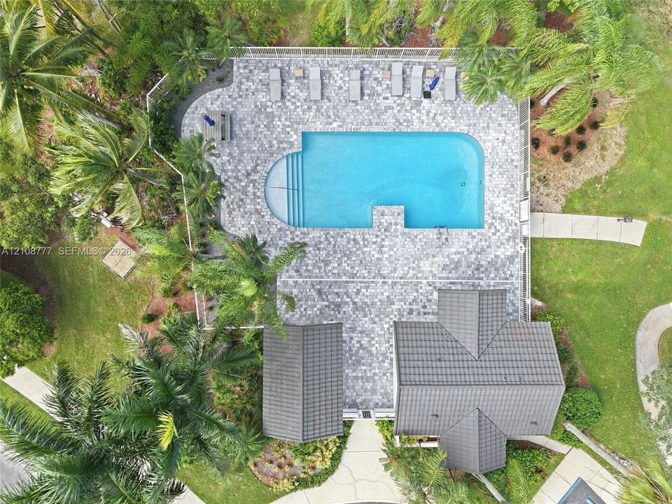 Aerial perspective of the grounds featuring a geometric swimming pool, extensive stone paver decking, enclosed perimeter fencing, and mature palm landscaping