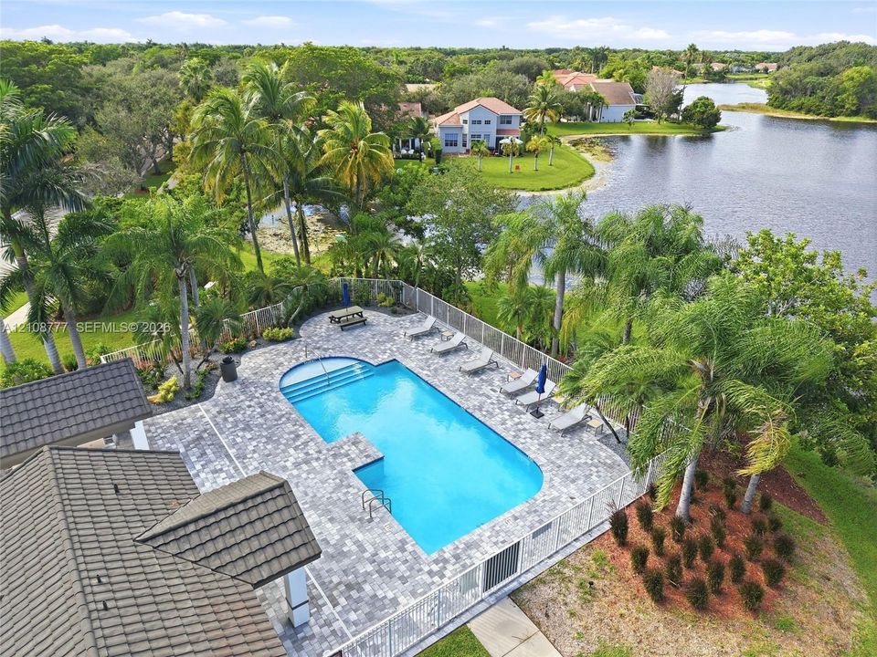 In-ground swimming pool with stone-paver decking, metal perimeter fencing, mature palm trees, and waterfront lot positioning