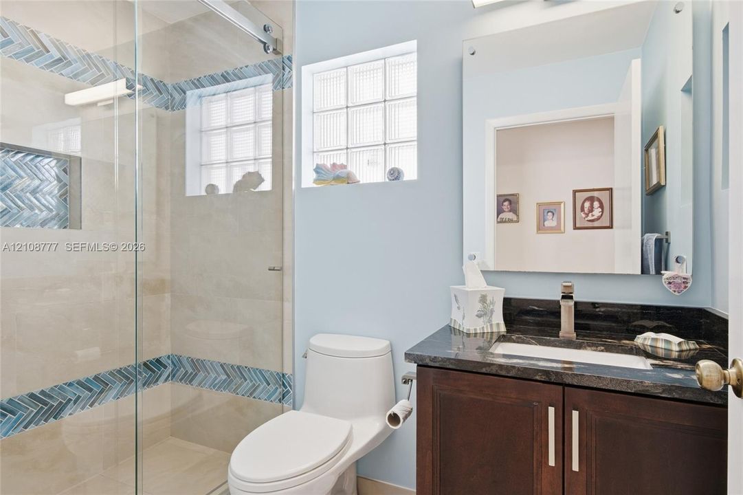 Full bathroom featuring a walk-in shower with glass sliding door and decorative herringbone-pattern tile inlay, a dark wood-finish vanity with a dark stone countertop, an integrated sink, and glass block windows