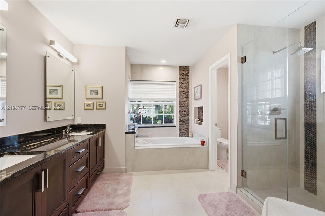 Double vanity with dark wood cabinetry and stone countertops, oversized walk-in glass shower with tile inlay, and built-in soaking tub