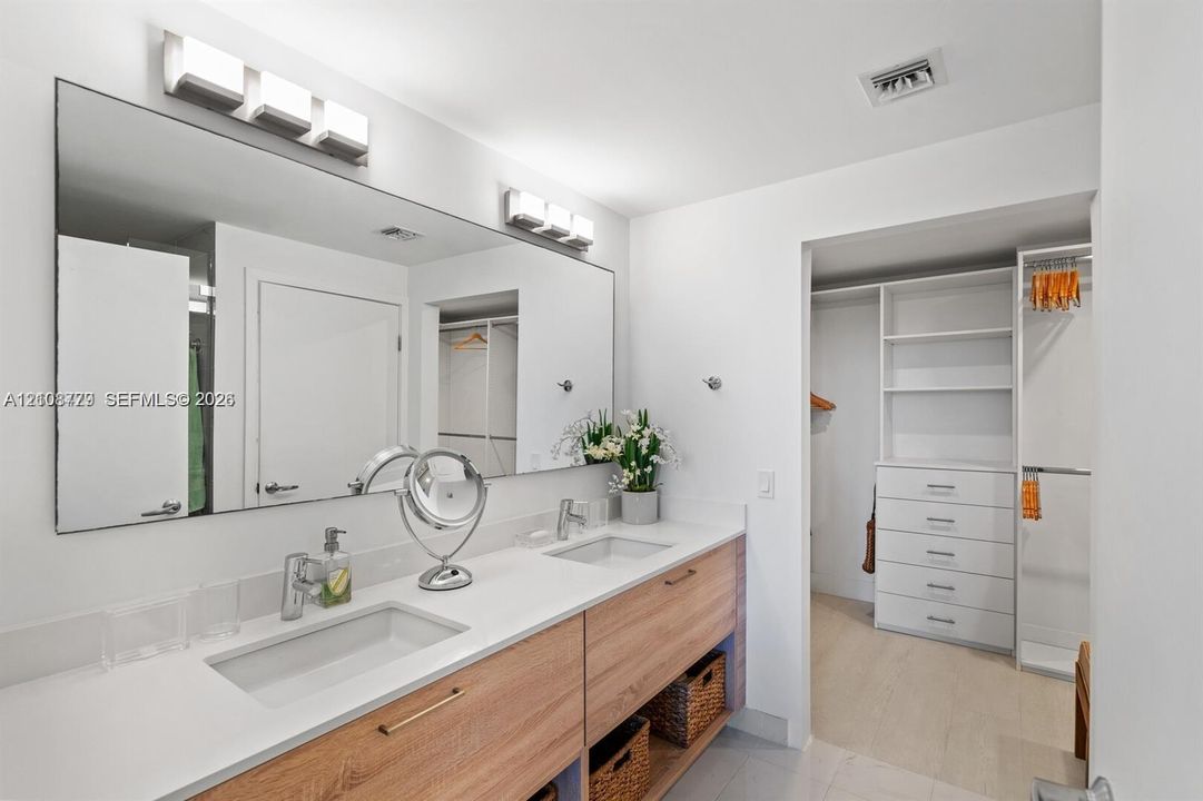 Dual-sink wood-finish vanity with a quartz countertop, full-width wall-mounted mirror, brushed metal fixtures, and walk-in closet featuring integrated shelving and drawer storage