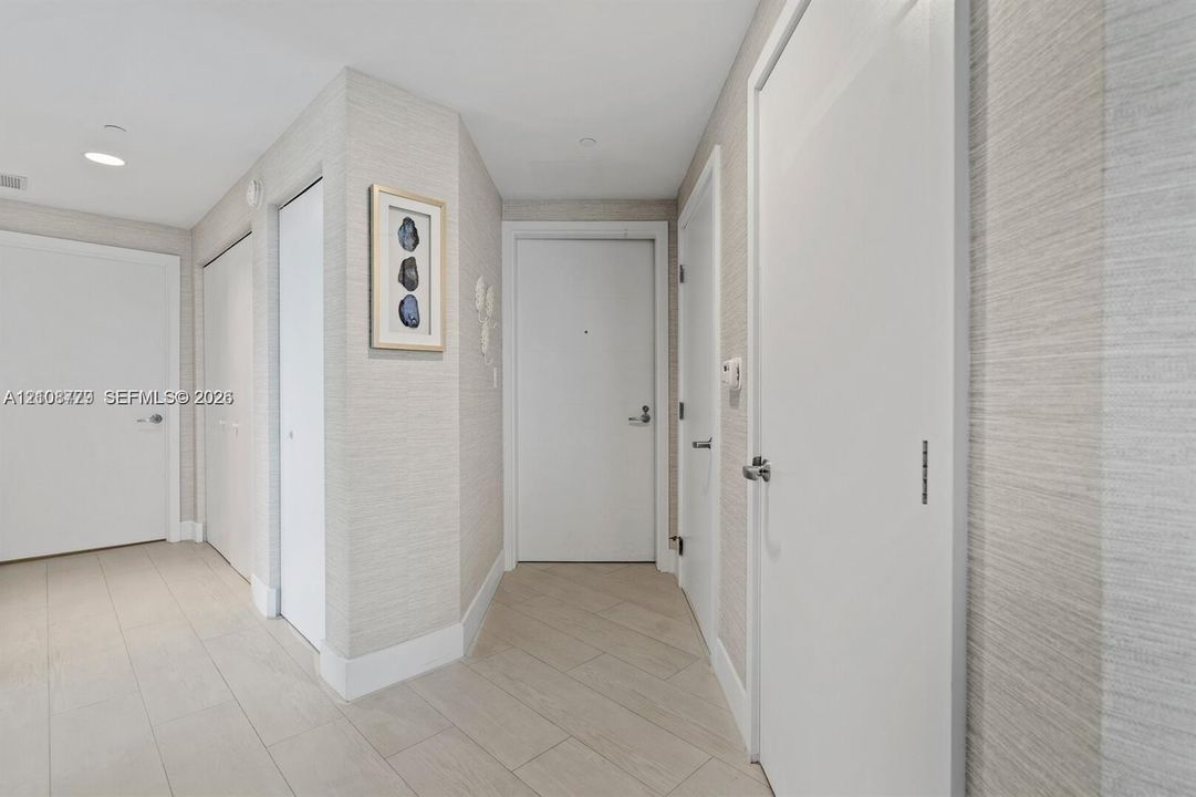 Entryway featuring neutral-toned textured wall covering, large-format tile flooring, multiple interior doors with brushed metal hardware, and recessed ceiling lighting
