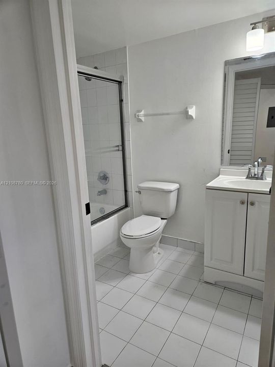 Bathroom featuring a white vanity cabinet with an integrated sink, a tiled tub-shower combination with a sliding glass door, white floor tiling, a framed wall mirror, and a wall-mounted light fixture