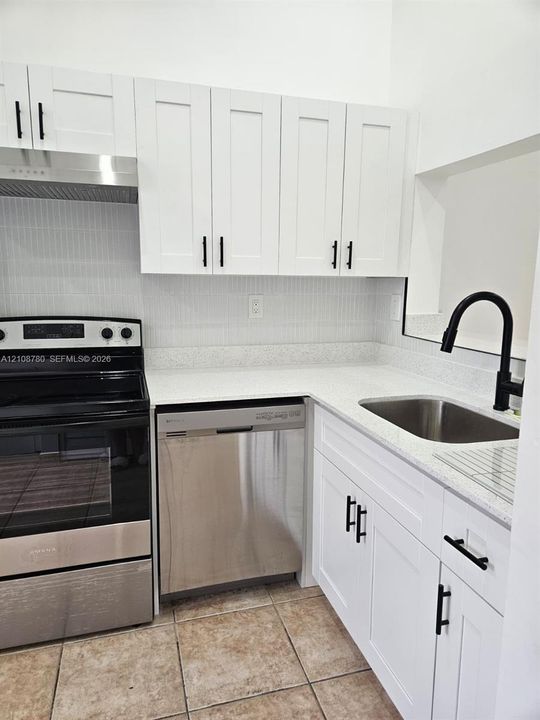 Kitchen featuring white shaker-style cabinetry, light-toned stone-composite countertops, stainless steel appliances, a matte black gooseneck faucet, and vertical-tile backsplash