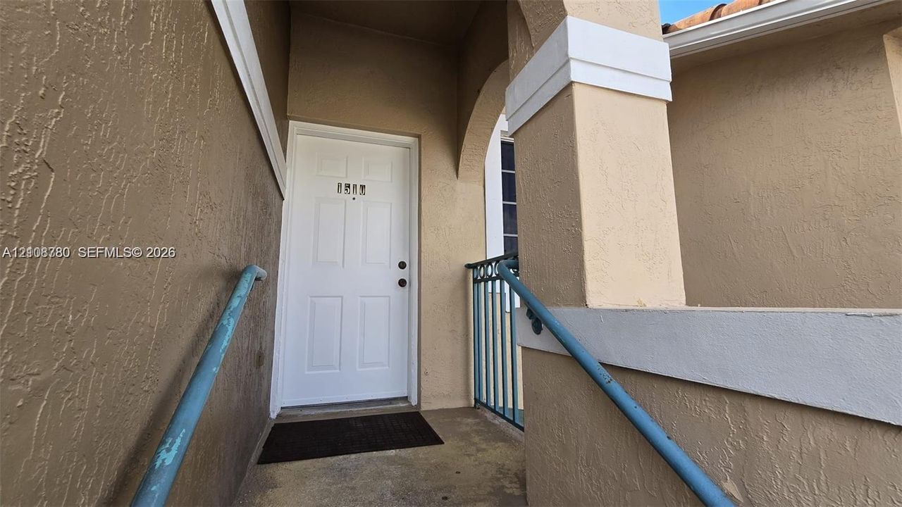 Stucco exterior entryway featuring a paneled door, arched architectural detailing, a stone-finish pillar, and blue metal railings