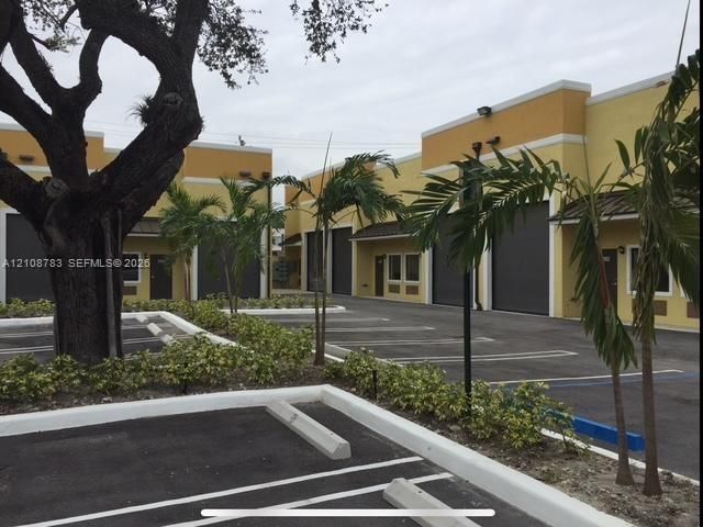 Commercial exterior featuring stucco facades, multiple roll-up doors, paved parking area with concrete curbing, and mature landscaping