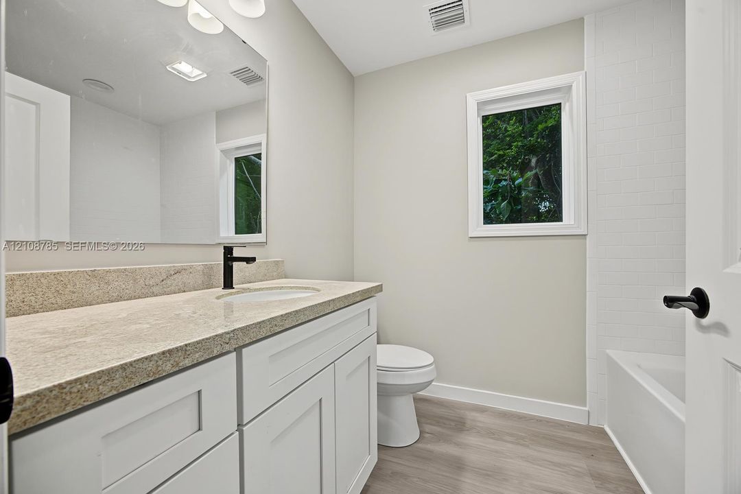 Bathroom featuring a white shaker-style vanity, stone-surface countertop, dark metal hardware, wood-finish flooring, and a subway-tile tub surround