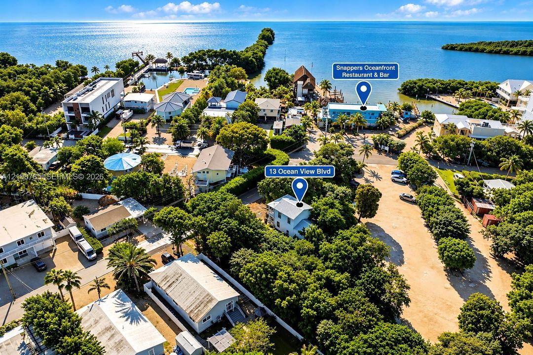 Aerial perspective of coastal residential property featuring mature landscaping, metal roofing, ground-level paved parking, and proximity to the open water