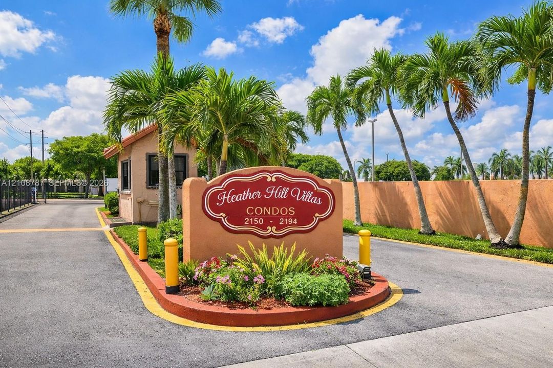 Gated community entrance featuring a masonry monument sign, professional landscaping, paved access roads, and mature palm trees