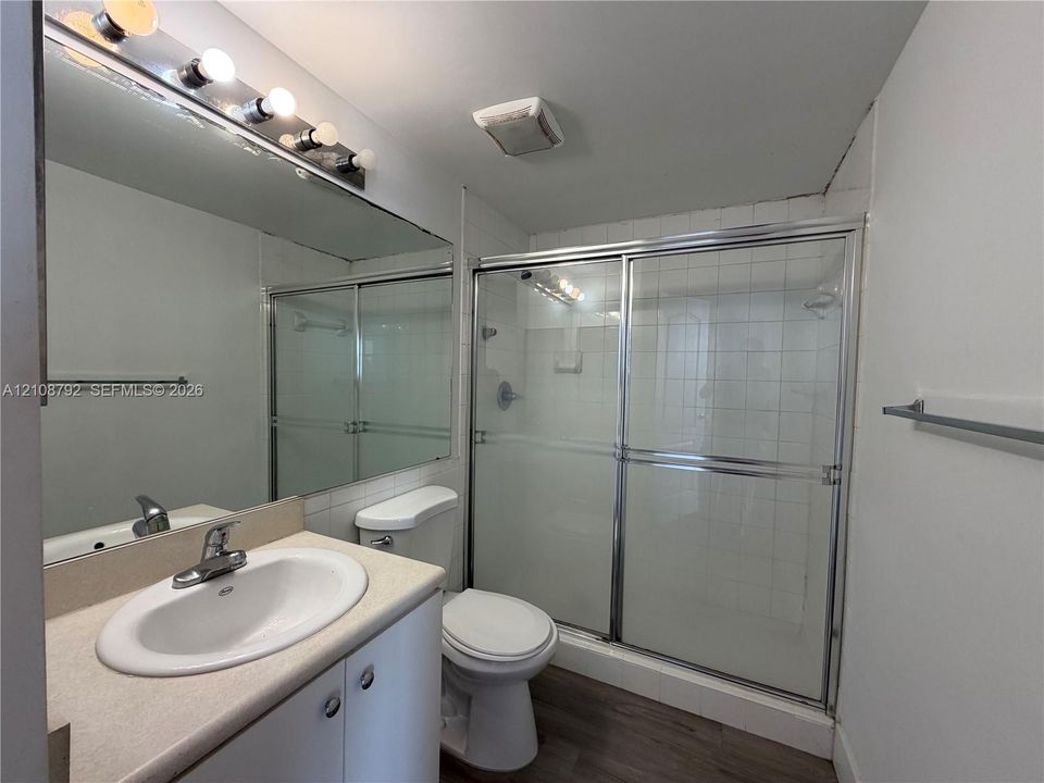 Bathroom featuring a vanity with an integrated basin, a large wall-mounted mirror with multi-bulb lighting, and a walk-in shower with sliding glass doors and tiled surround
