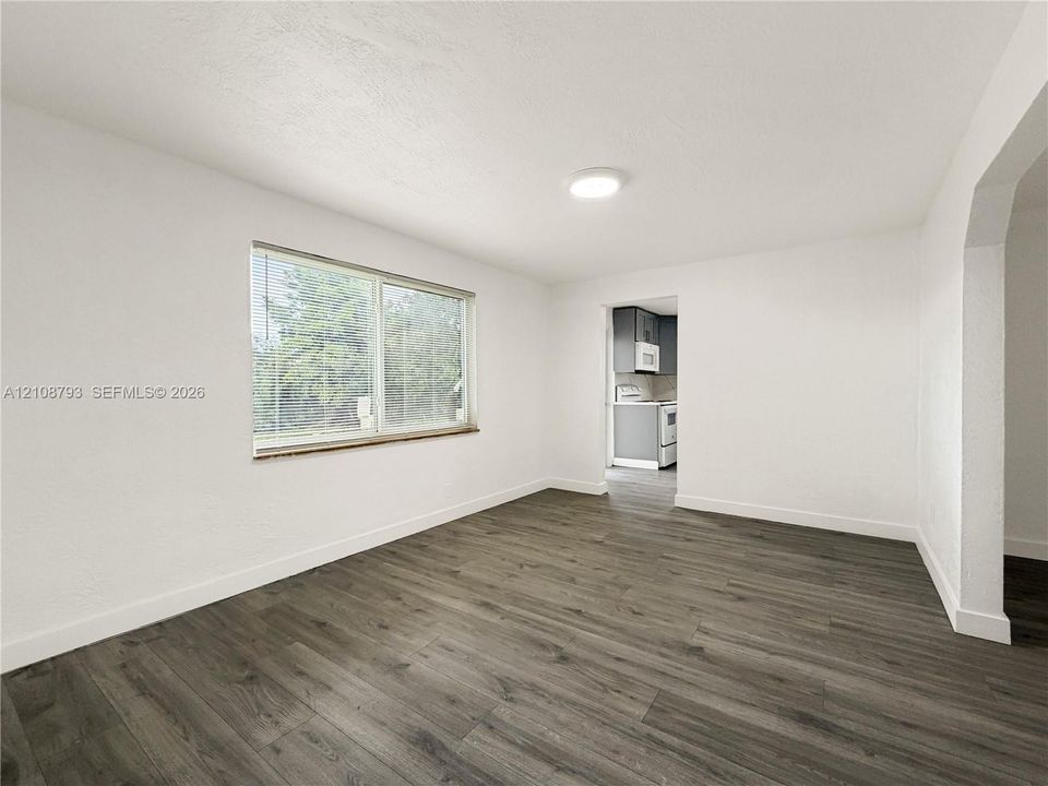 Neutral-toned interior featuring wood-finish flooring, white walls, flush-mount ceiling lighting, a large window with horizontal blinds, and a wide arched opening