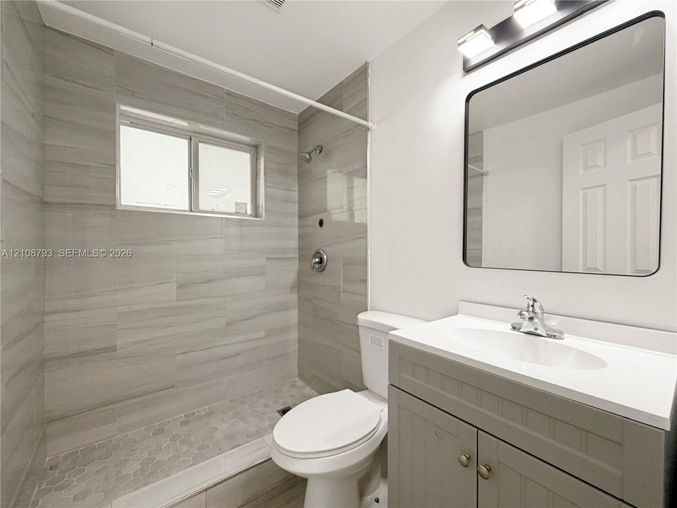 Walk-in shower featuring large-format tile walls, integrated hexagonal floor tiling, grey cabinetry with a white countertop, and a wall-mounted rectangular mirror
