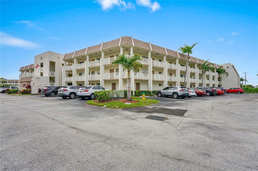 Multi-story residential building featuring a beige stucco exterior, tiered open-air walkways with white railings, and paved surface parking