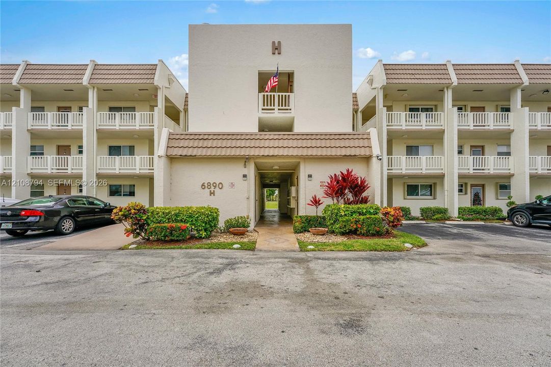 Multi-story building exterior featuring a neutral stucco facade, tiered balconies with white railings, tile roofing, and curated shrubbery