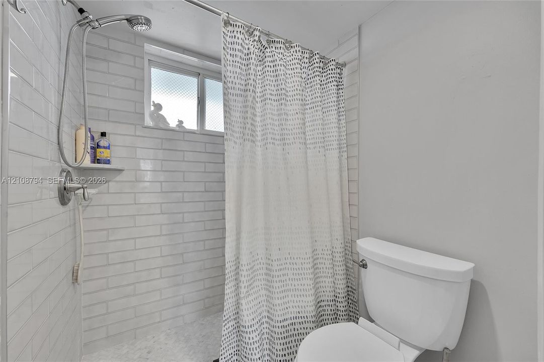 Bathroom featuring horizontal white subway tile shower surround, adjustable handheld shower fixture, built-in corner shelving, and white high-profile toilet
