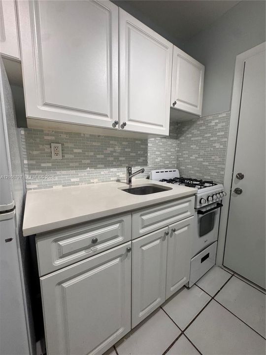 Kitchen featuring white shaker-style cabinetry, solid surface countertops, a mosaic tile backsplash, a stainless steel undermount sink, and ceramic tile flooring