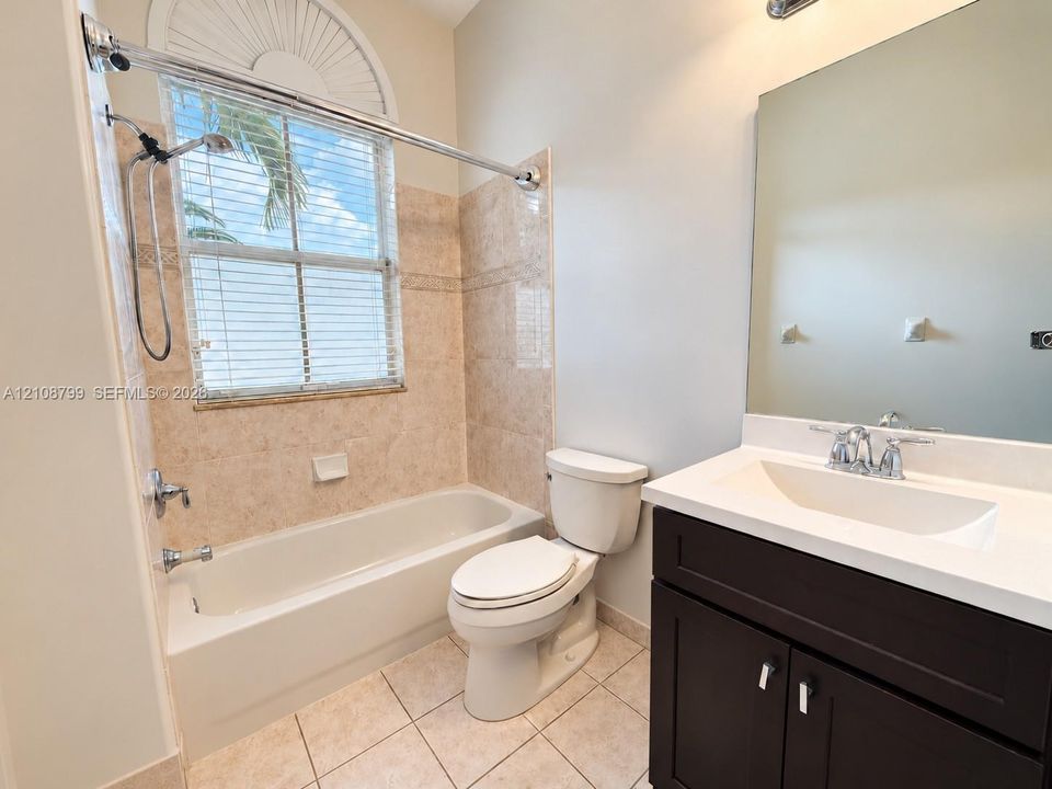Bathroom featuring a dark wood-finish vanity with a white countertop and integrated sink, a tiled tub-shower combination, ceramic tile flooring, and a window with an arched transom