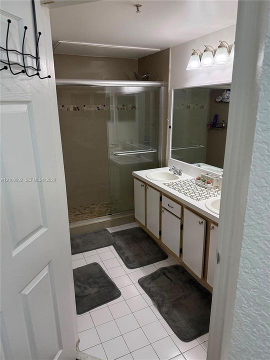Bathroom featuring a dual-sink vanity with white cabinetry, a large wall-mounted mirror, a tiled shower enclosure with sliding glass doors, and white ceramic tile flooring