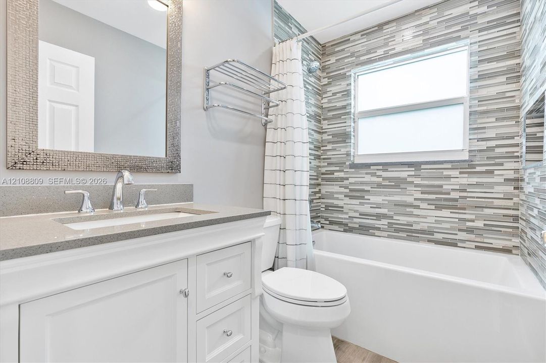 Bathroom featuring a white vanity cabinet with a stone-composite countertop, rectangular undermount sink, mosaic-tiled tub surround, and a wall-mounted metal shelving unit