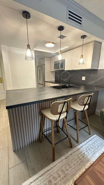 Kitchen featuring a peninsula with a dark countertop and slatted base, wood-finish flooring, three suspended pendant lights, and dark stone-finish backsplash