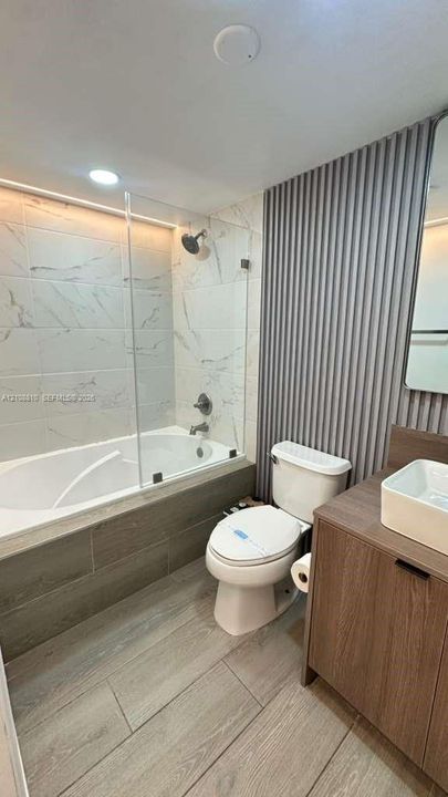 Bathroom featuring a wood-finish vanity with a vessel sink, a glass-enclosed soaking tub with stone-finish surround, vertical-slat wall paneling, and wood-finish flooring