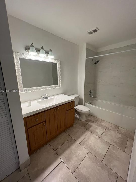Bathroom featuring wood-finish cabinetry, a solid-surface countertop with an integrated rectangular sink, large-format tile flooring, and a bathtub surround with tiled walls