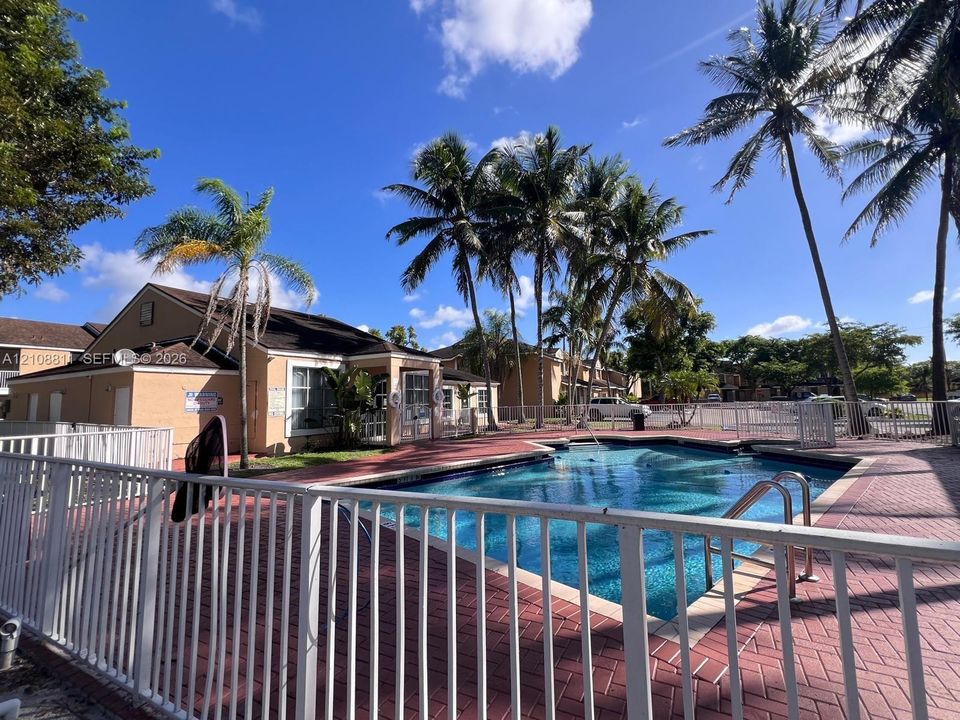 In-ground swimming pool featuring a herringbone-patterned brick paver deck, white metal perimeter fencing, and mature palm trees